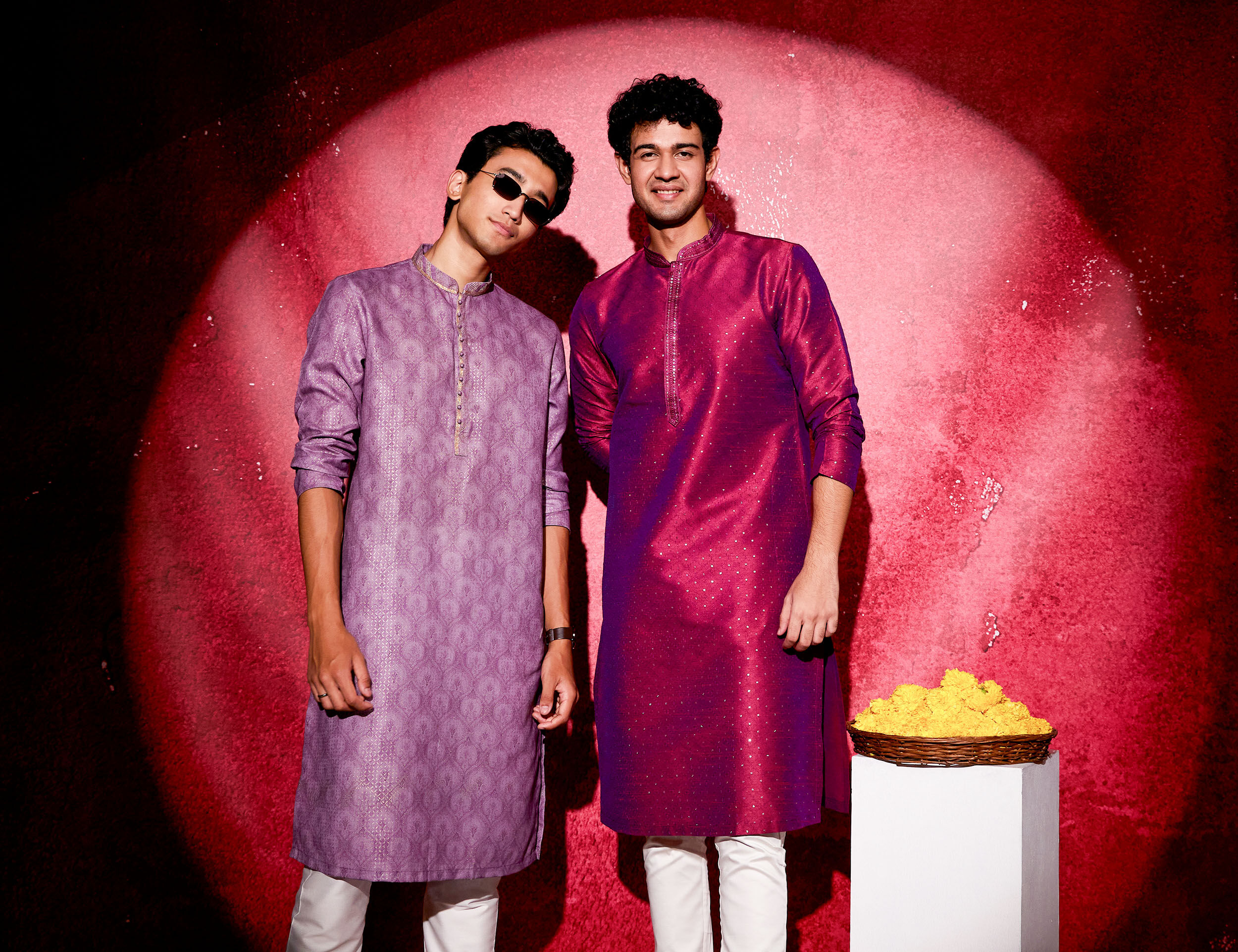 Two men in festive casual wear, one in lavender kurta and the other in deep magenta kurta, styled with white pants against a red backdrop with marigold flowers